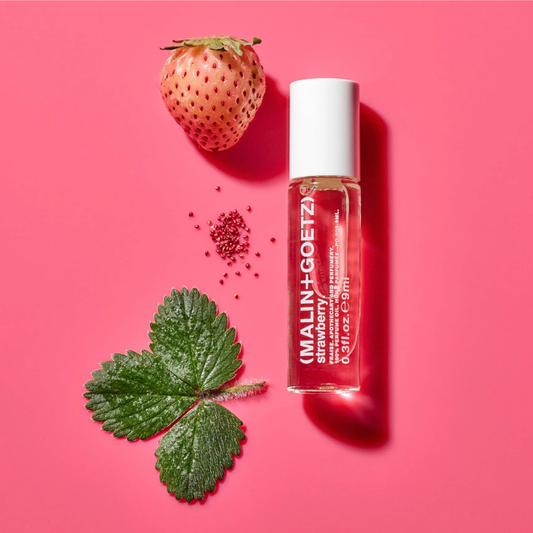 A clear cylindrical roller bottle labeled MALIN+GOETZ Strawberry & Rosehip Infused Serum, styled on a bright pink background with a whole strawberry, a strawberry leaf, and small red seed pieces nearby.