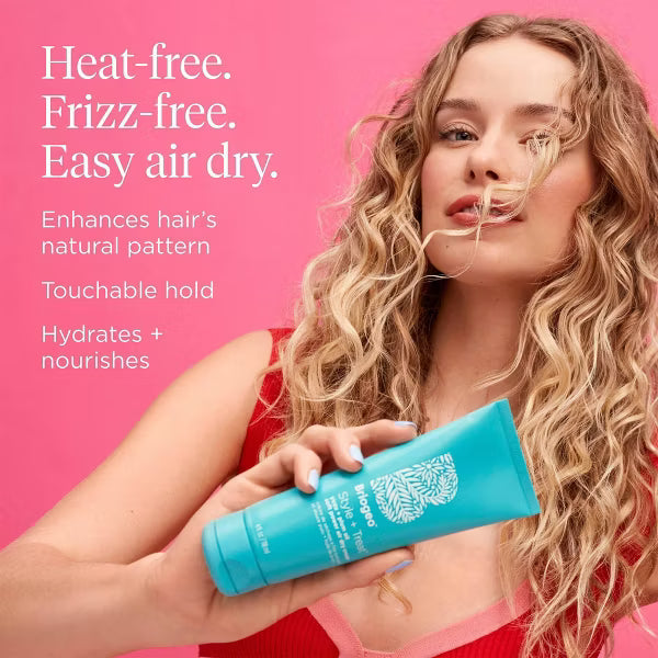 Model with long blonde waves holding Briogeo Soft Power Air Dry Cream against a pink background, highlighting heat-free styling and touchable hold.