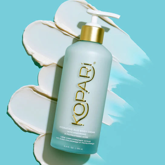 Kopari Hydrating Silk Body Cream bottle displayed over swirls of cream on a blue background