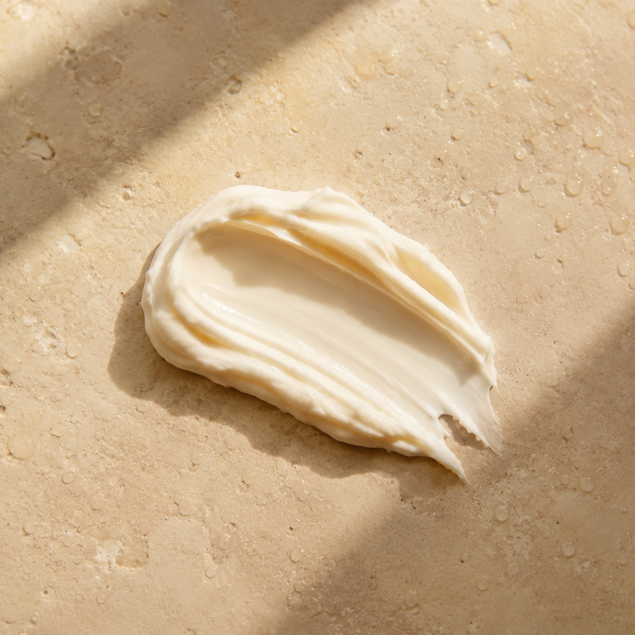 Ivory cream body cleanser texture on warm beige stone in soft sunlight