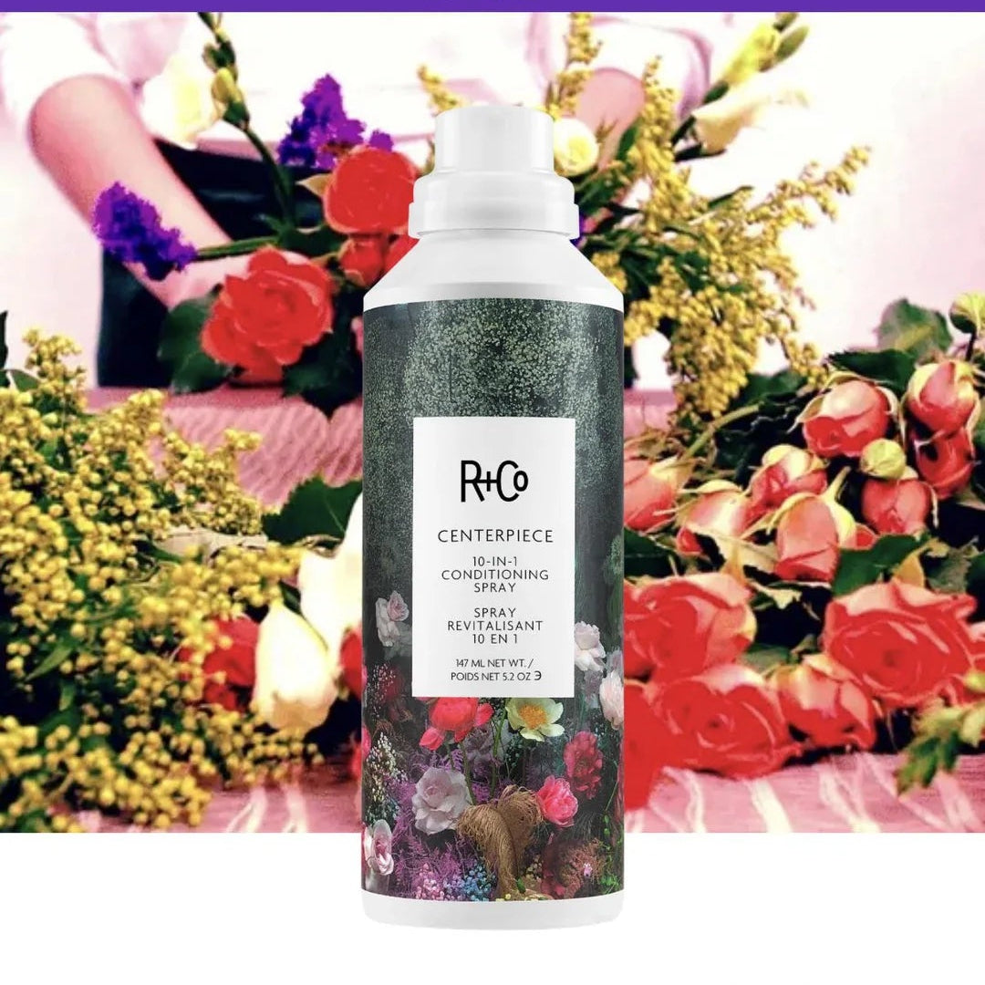 R+Co CENTERPIECE 10-IN-1 Conditioning Spray full-size bottle against a colorful floral backdrop.