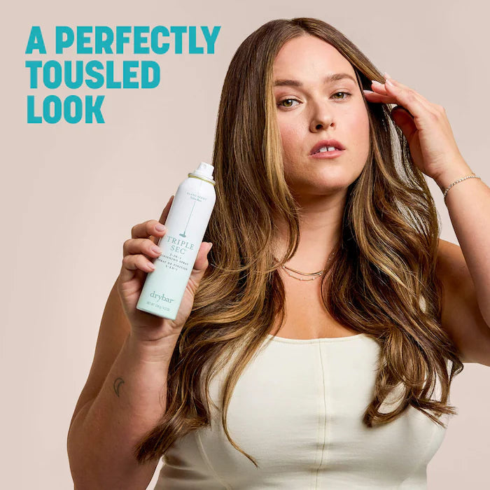 Model holding Drybar Triple Sec with long tousled waves against a blush background